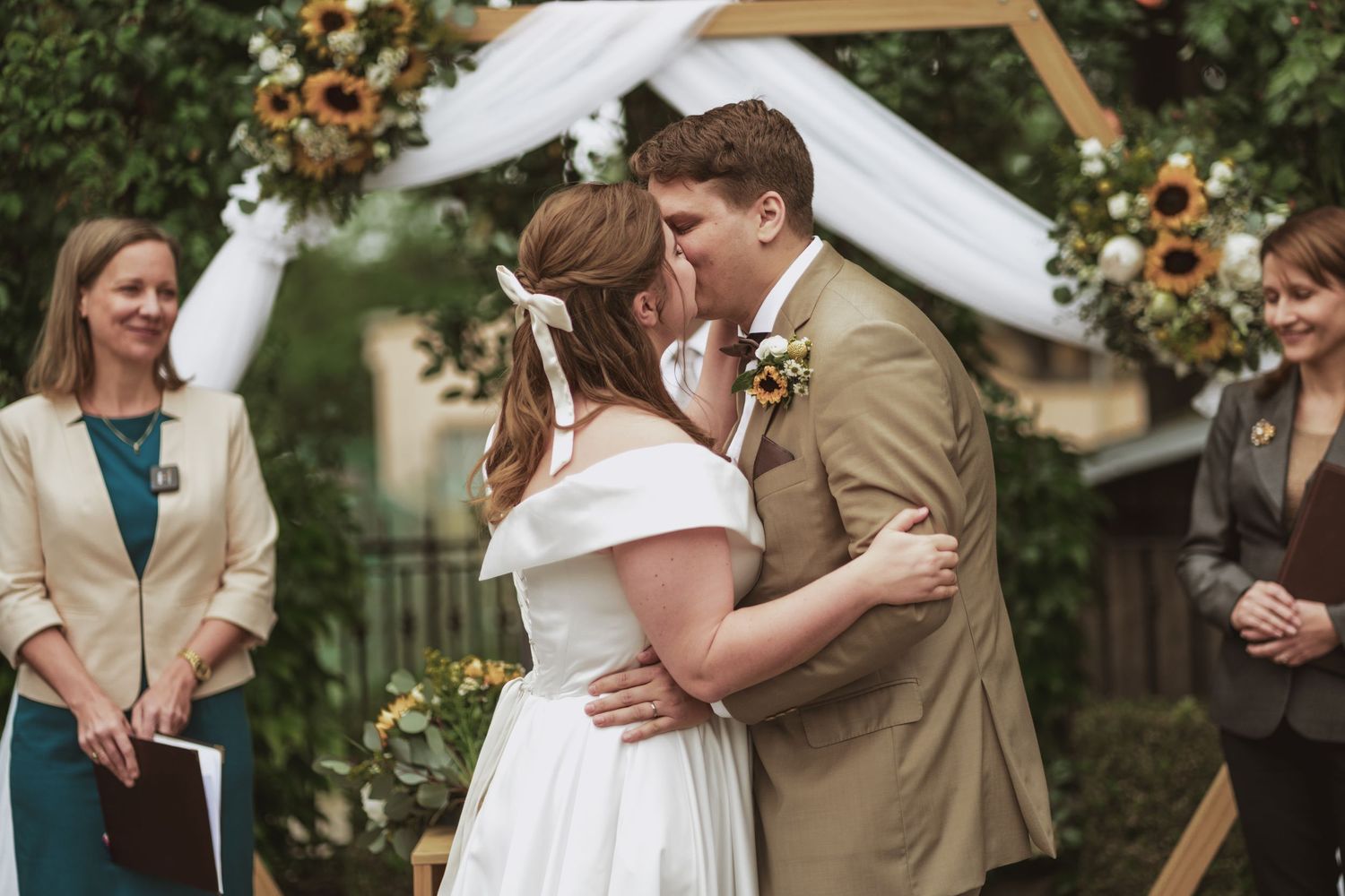 Bride in white gown and groom in tan suit kiss under a floral wedding arch with sunflowers as the officiant and a guest smile nearby.