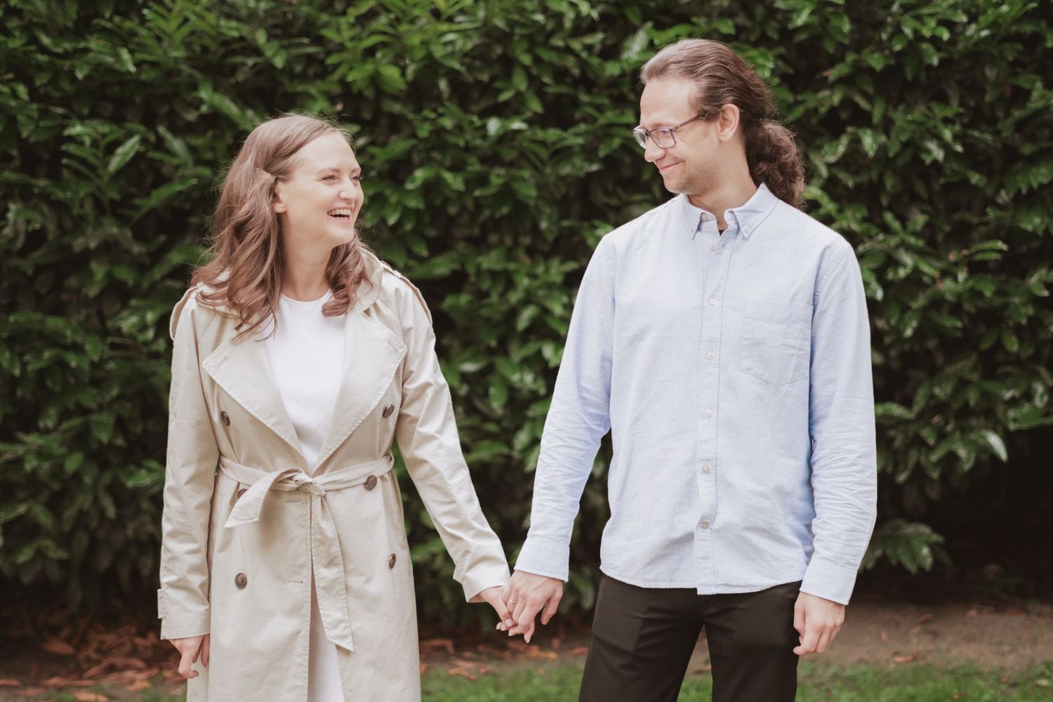 Smiling couple holding hands and laughing together outdoors, the woman in a beige trench coat and the man in a light blue shirt.