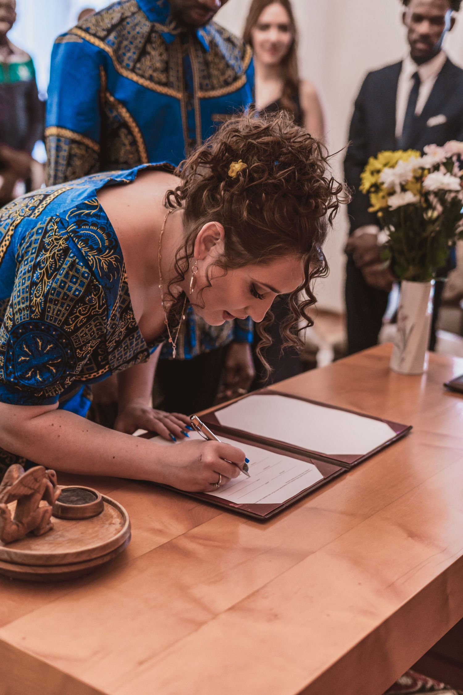 Woman in a blue patterned dress signs a document at a wooden table during a ceremony, with guests and a flower vase behind her.