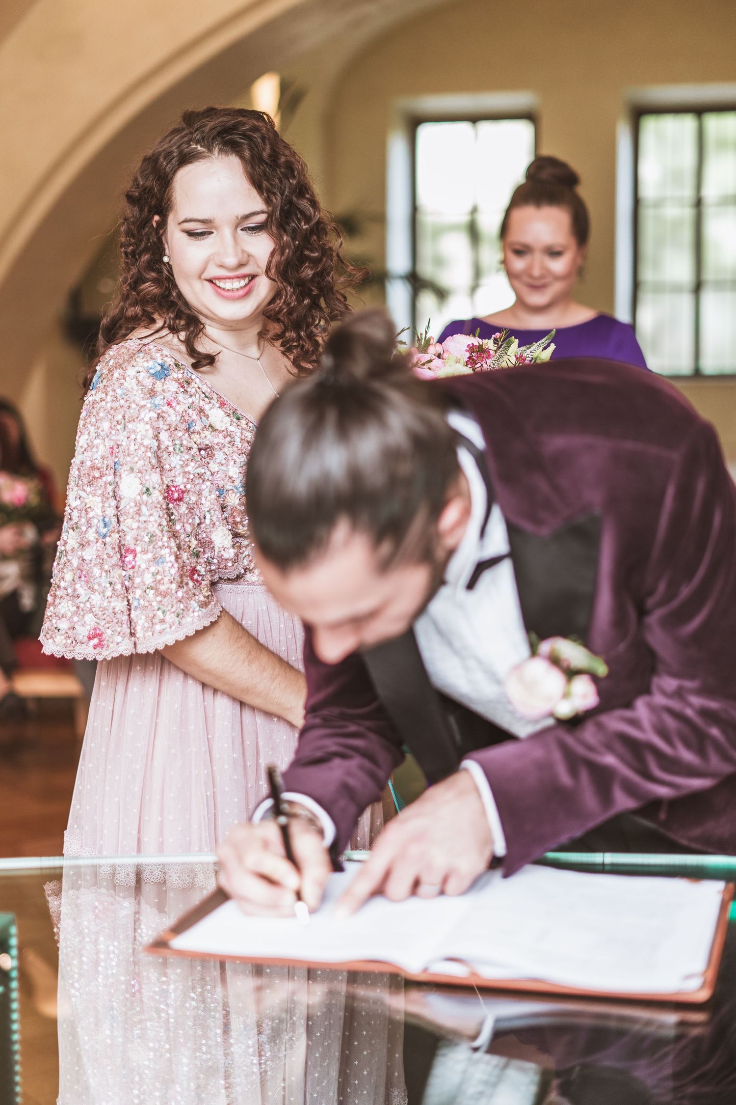 Smiling bride watches her groom in a burgundy velvet jacket sign the marriage register while a guest looks on behind them.