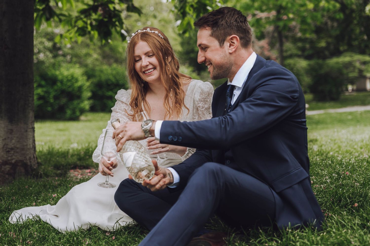 Smiling bride in a white dress and groom in a navy suit sit together on green grass in a sunny park, sharing a joyful wedding moment.