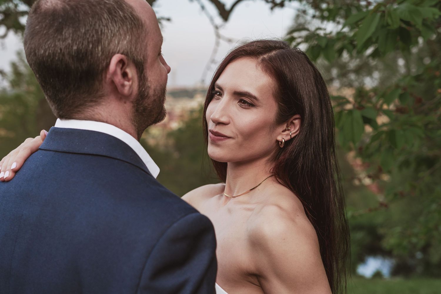 Bride in an off-the-shoulder dress with long dark hair gazes at a bearded groom in a navy suit, outdoors with green trees behind.