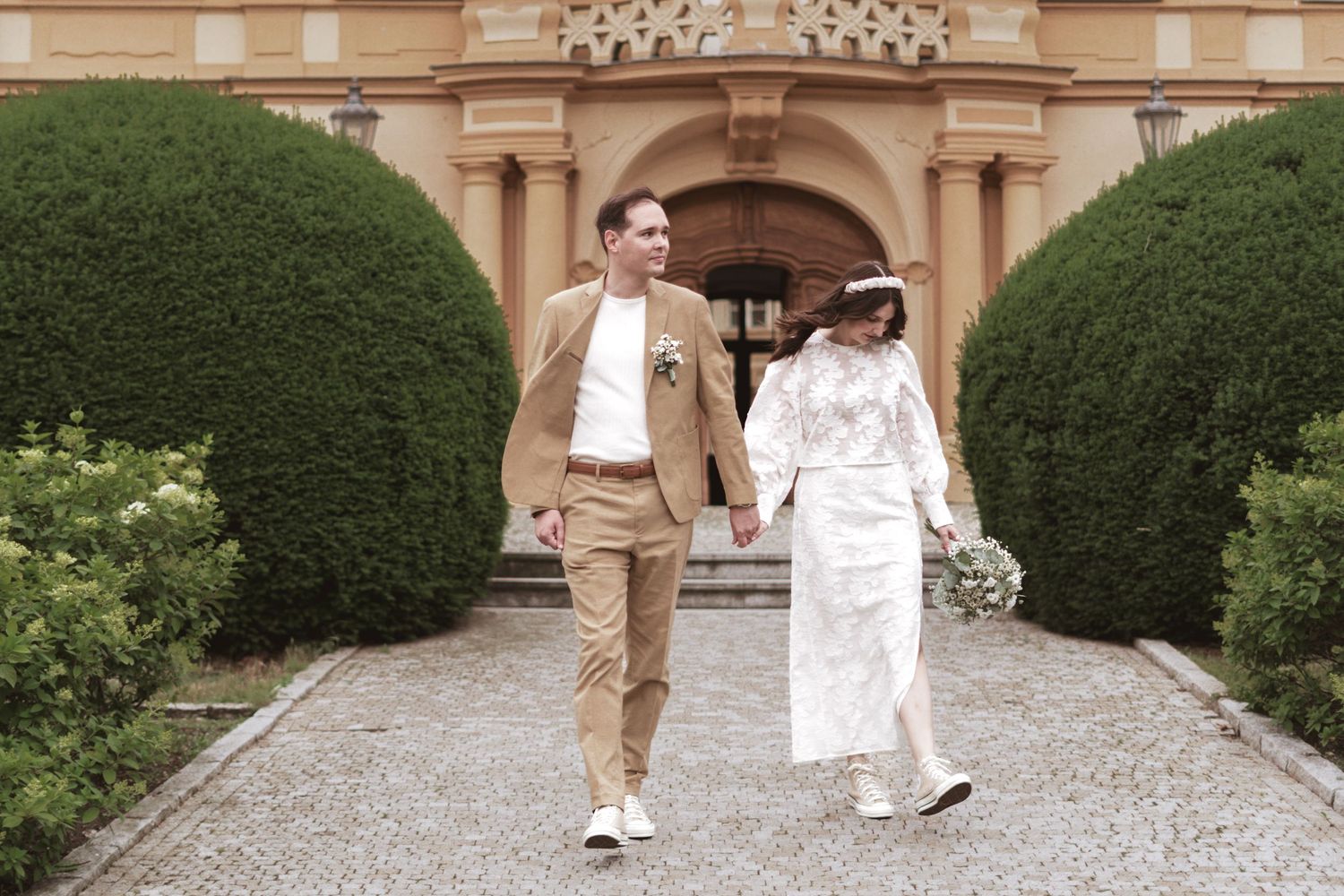 Bride in a white dress and groom in a tan suit hold hands while walking down a stone path past trimmed hedges toward a historic building.