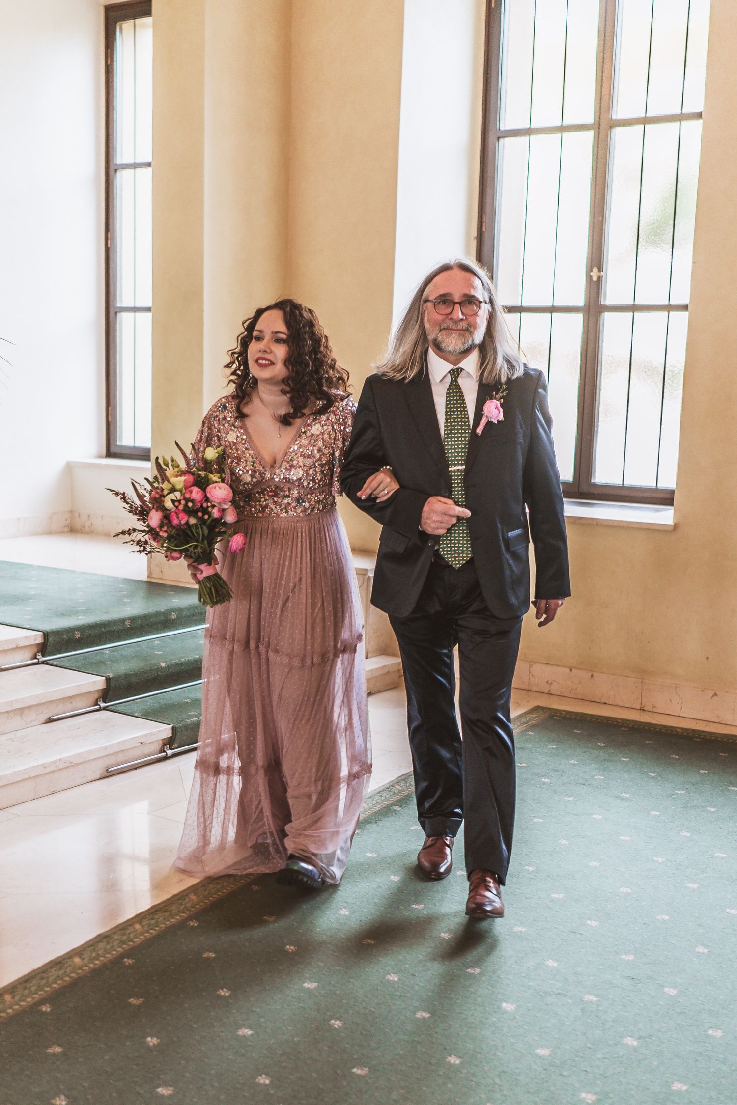 Bride in a blush pink sequined gown holding a bouquet walks arm in arm with a long-haired man in a dark suit down a green-carpeted hall.