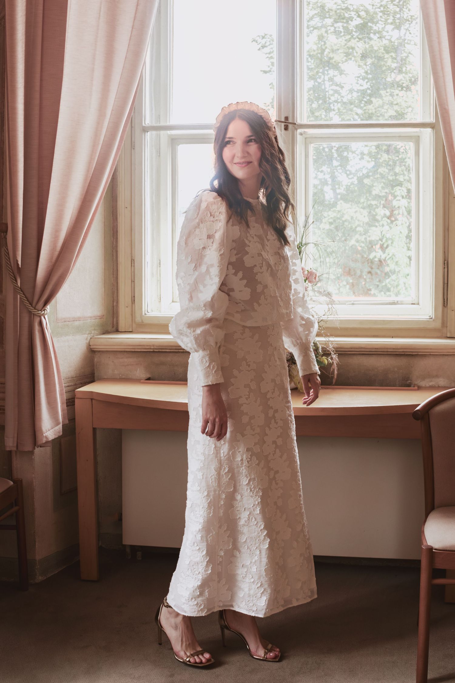Woman in an ivory lace skirt set with puff sleeves and strappy heels stands beside a sunlit window and a wooden table.