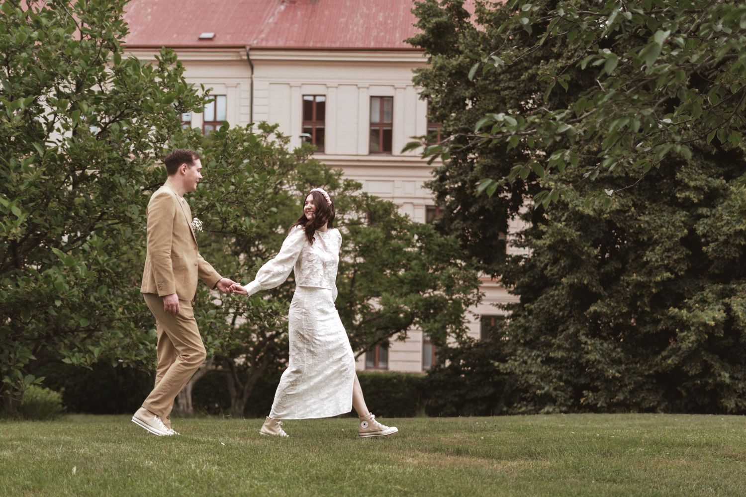 A man in a tan suit and a woman in a white dress hold hands while walking across a green lawn near trees and a historic building.