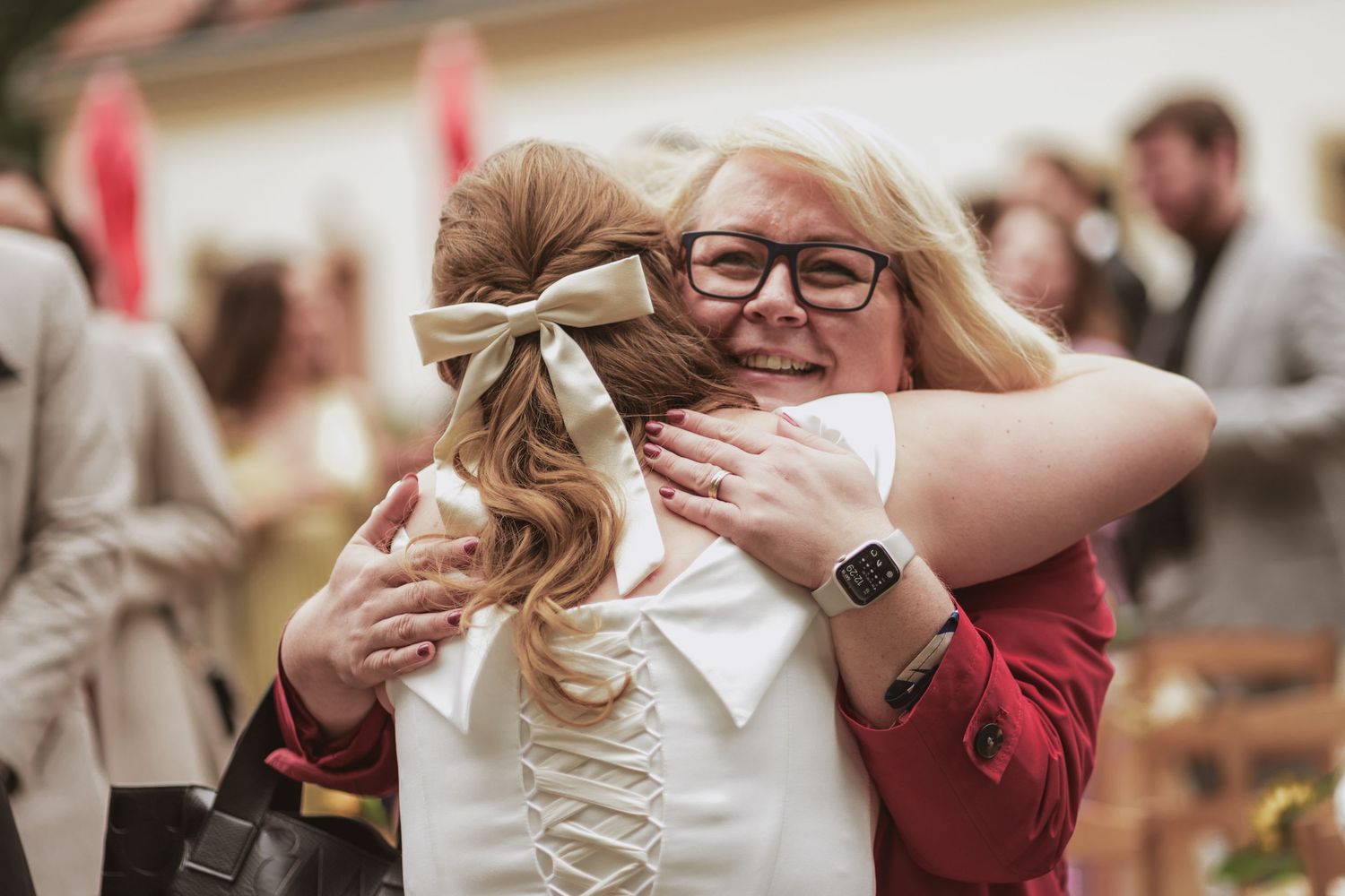 Smiling woman with glasses in a red cardigan hugs a woman wearing a white lace-up top and tan hair bow at a lively outdoor gathering.