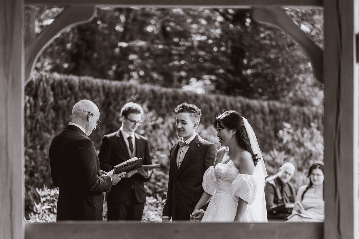 Black and white photo of a bride and groom laughing during an outdoor wedding ceremony as an officiant reads and a guest looks on.