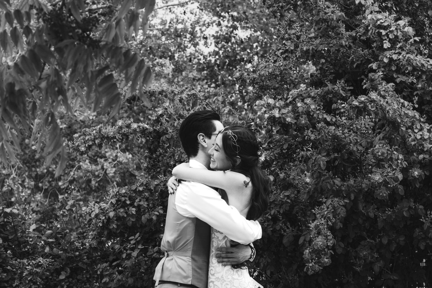 Black and white photo of a couple embracing and kissing outdoors in front of lush green trees, the groom wearing a vest and glasses.