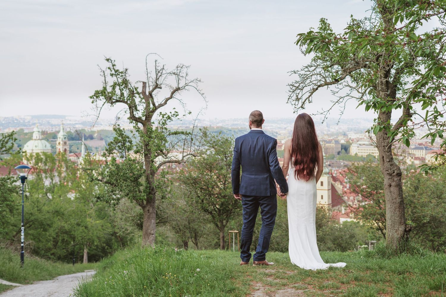 Bride in a white gown and groom in a navy suit hold hands on a grassy hilltop overlooking a scenic European city skyline.