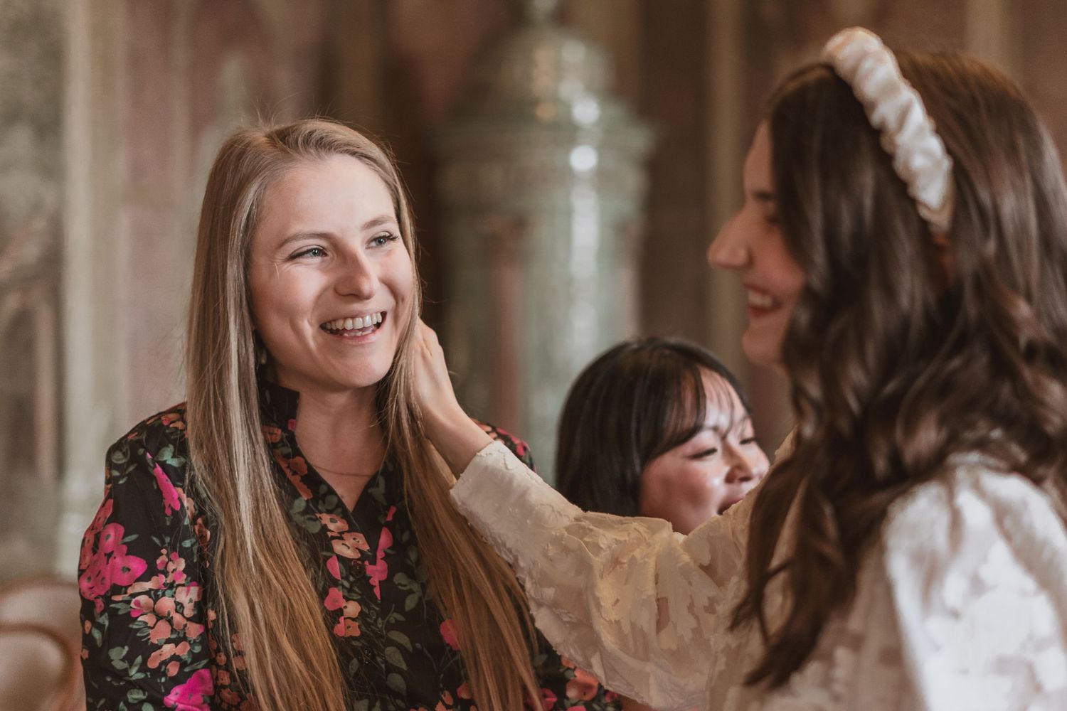 Smiling woman with long blonde hair in a floral blouse laughs as another woman in a white lace top and headband touches her face.