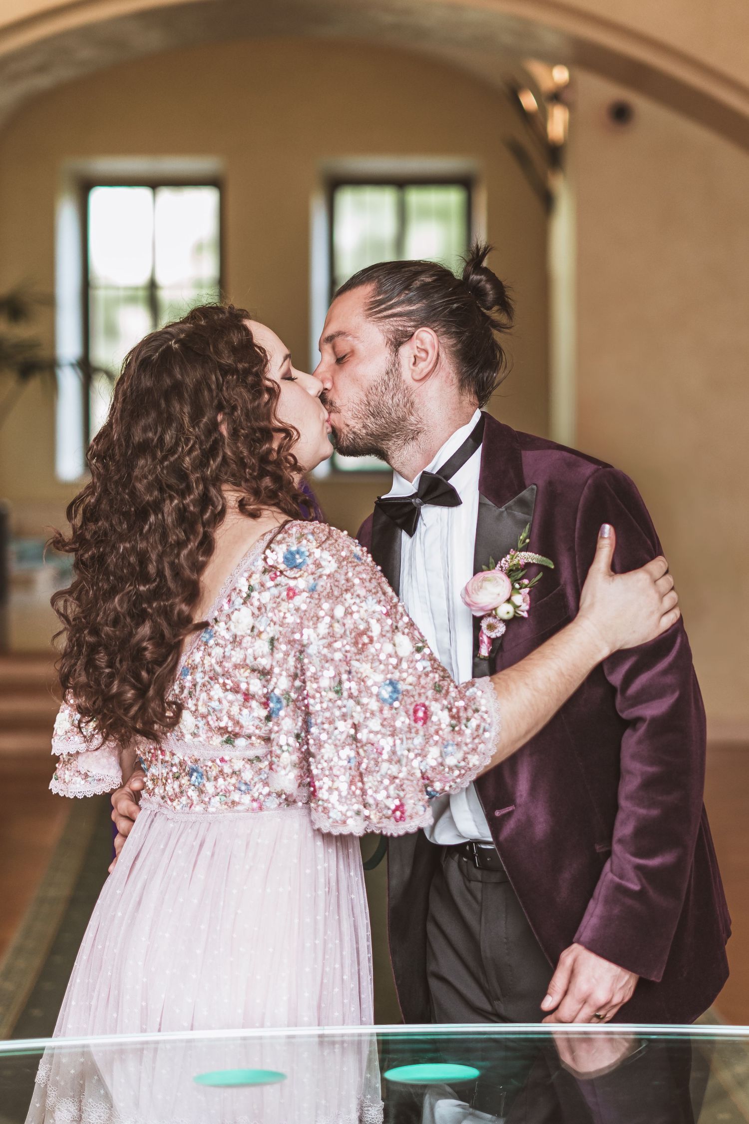 Bride in a pink beaded gown and groom in a burgundy velvet jacket share a wedding kiss inside a bright, arched venue.