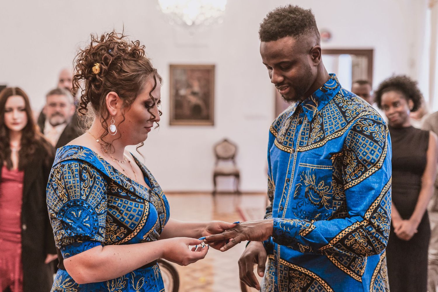 Couple in matching blue African print outfits exchange rings during a wedding ceremony in an elegant hall with guests looking on.