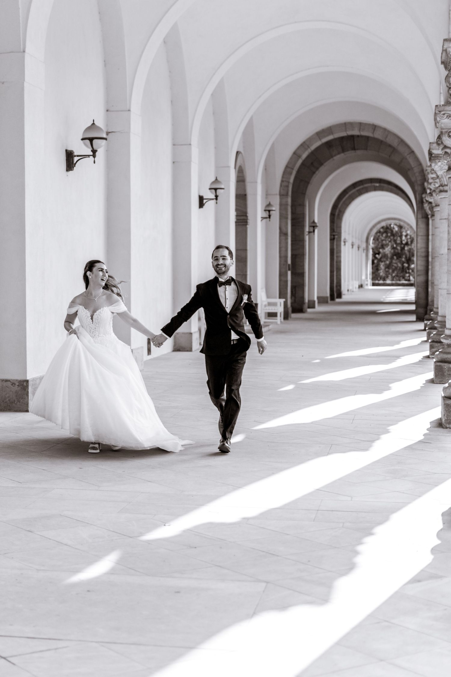 Bride in a white gown and groom in a tuxedo hold hands and walk through a sunlit arched corridor in a black and white wedding photo.
