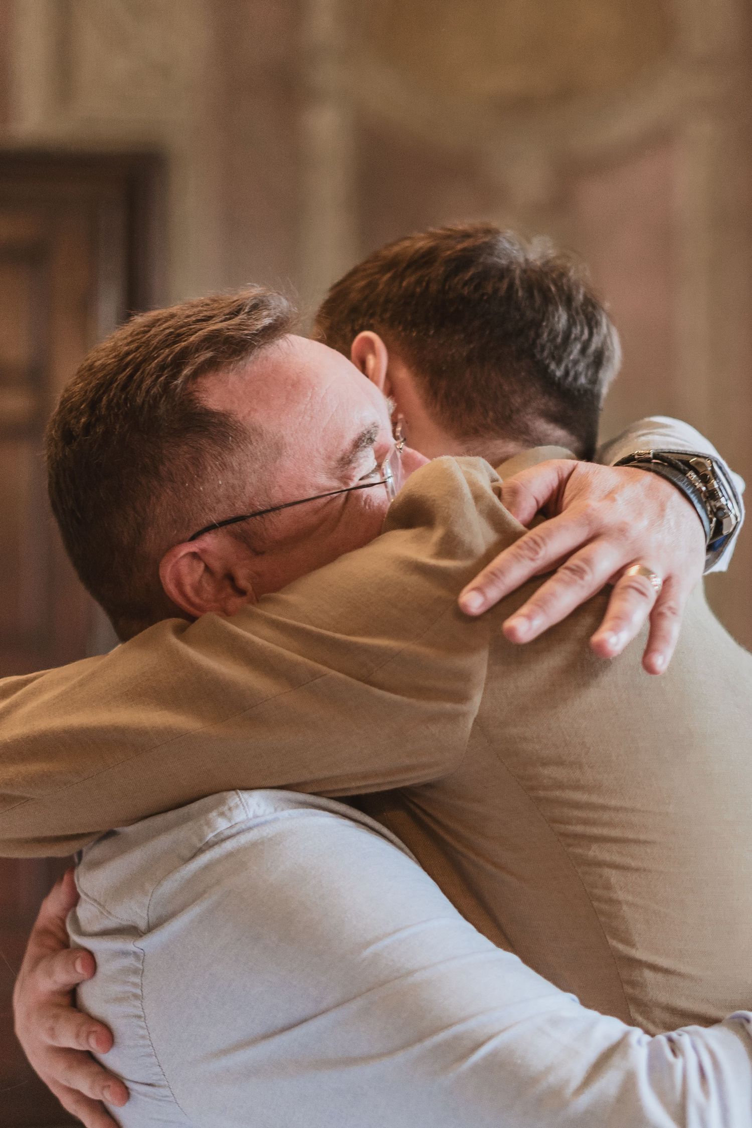 Two men in tan and light blue shirts share a warm, emotional hug inside an elegant room with ornate wooden doors.