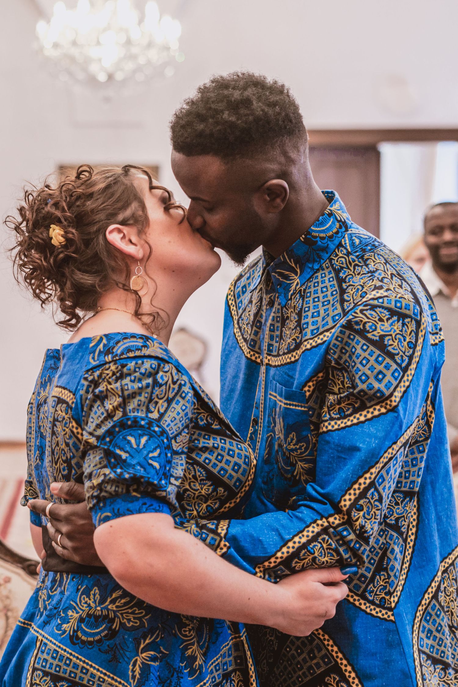 A couple in matching blue and gold African print outfits share a kiss during a wedding ceremony in an elegant room with a chandelier.