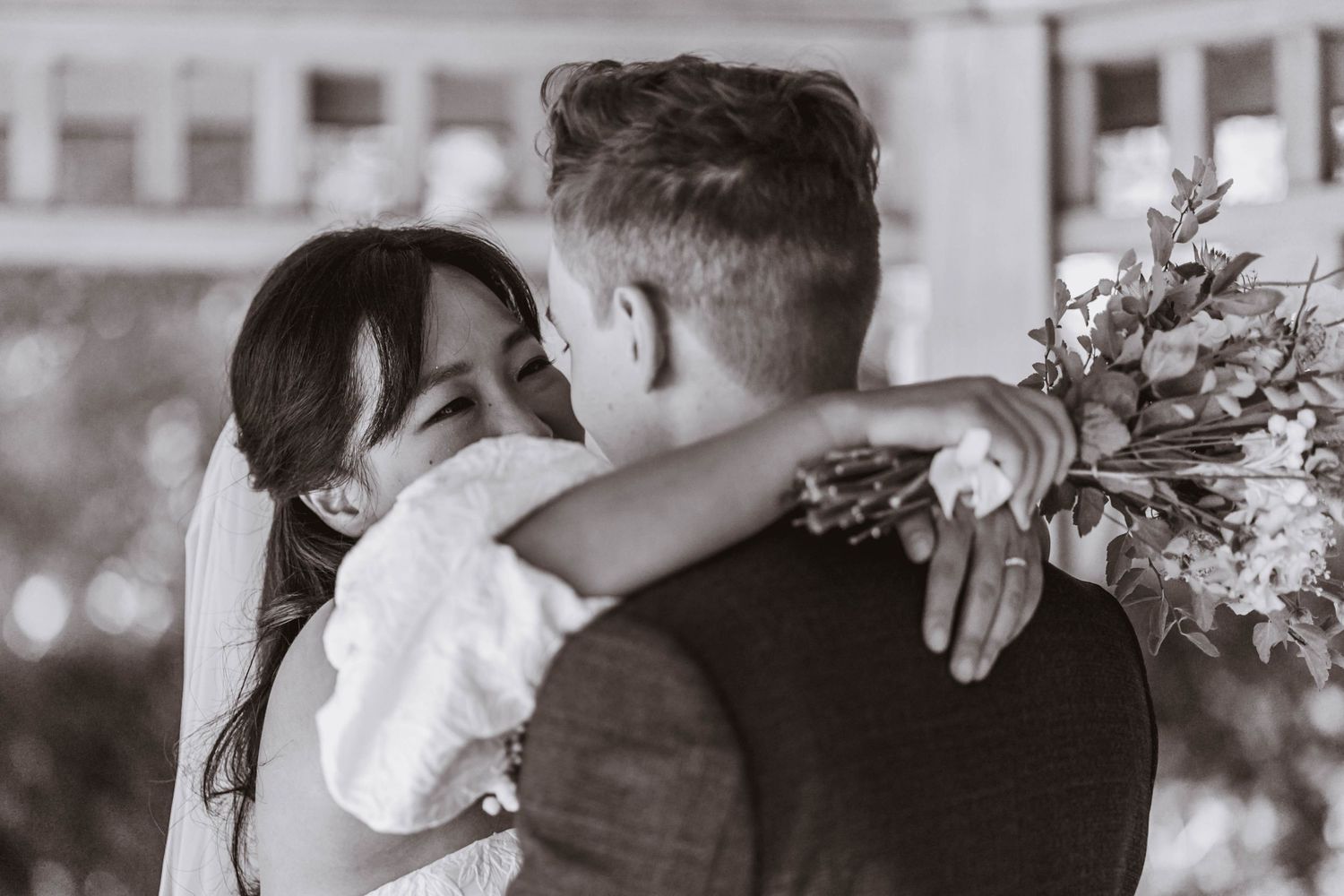Black and white photo of a smiling bride in a veil embracing her groom while holding a floral bouquet at an outdoor wedding.