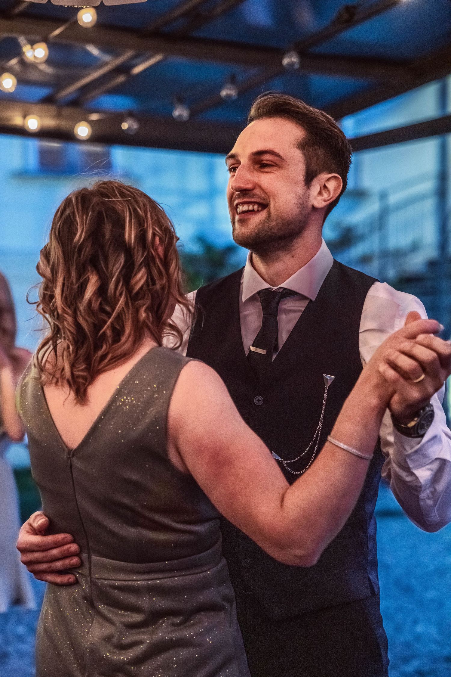 A smiling man in a black vest and tie dances with a woman in a gray dress under string lights at an evening wedding reception.