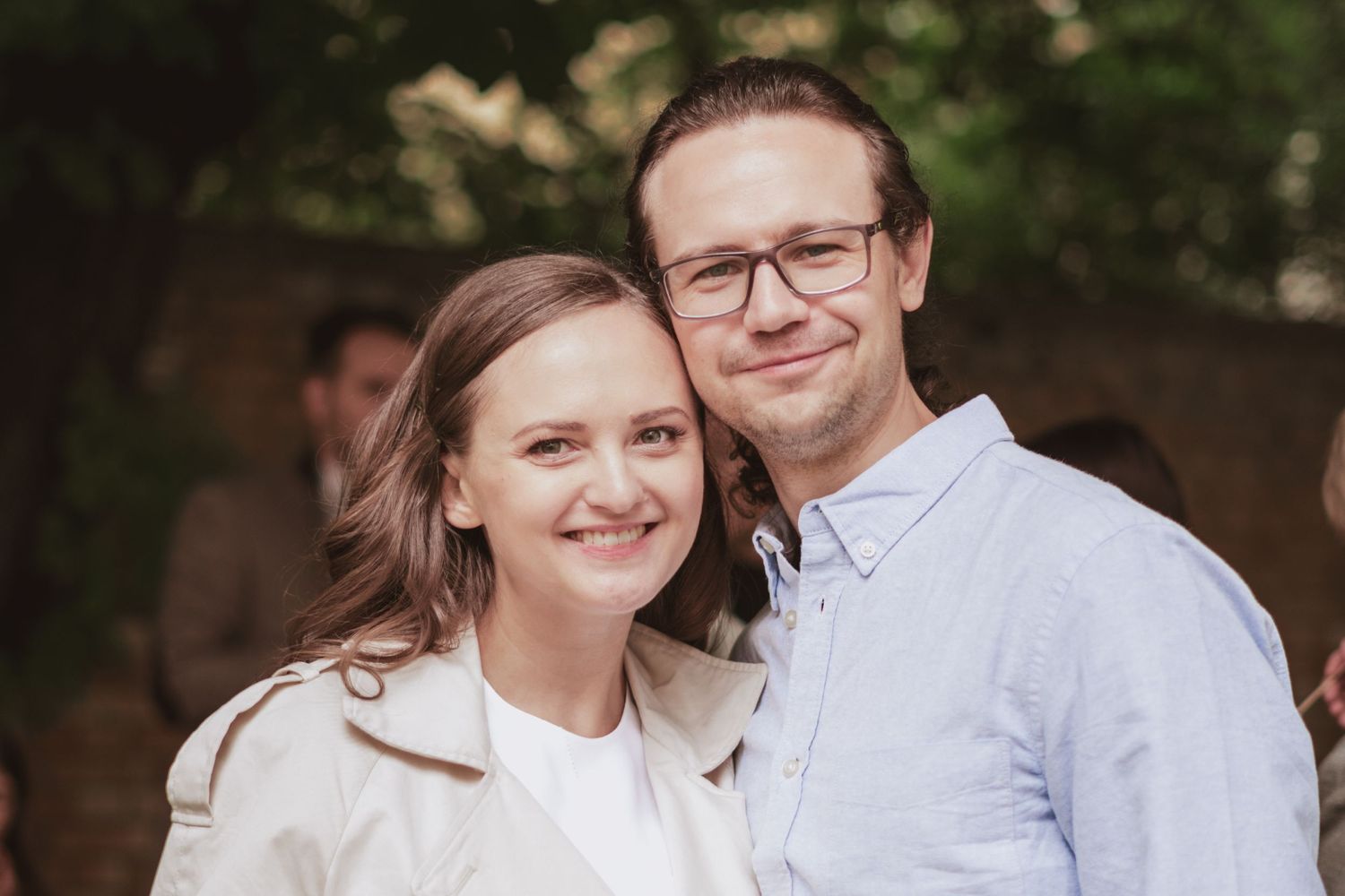 Smiling woman in a beige trench coat and a bespectacled man in a light blue shirt pose close together at an outdoor gathering.