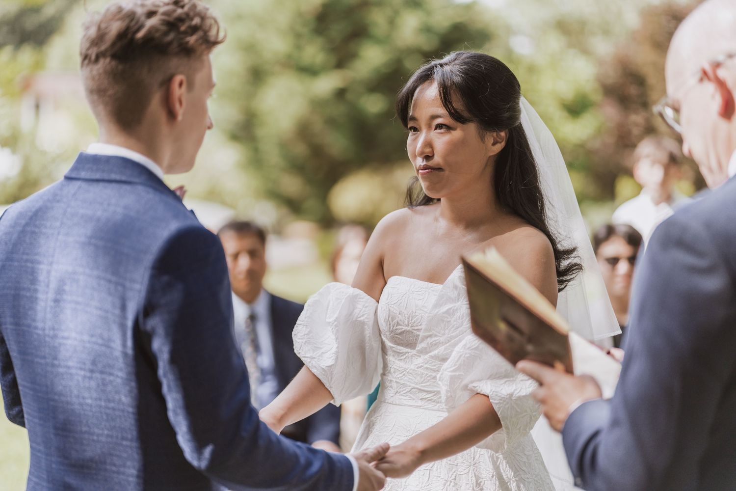 Bride in a white off-the-shoulder gown and veil holds hands with the groom during an outdoor wedding ceremony as an officiant reads.
