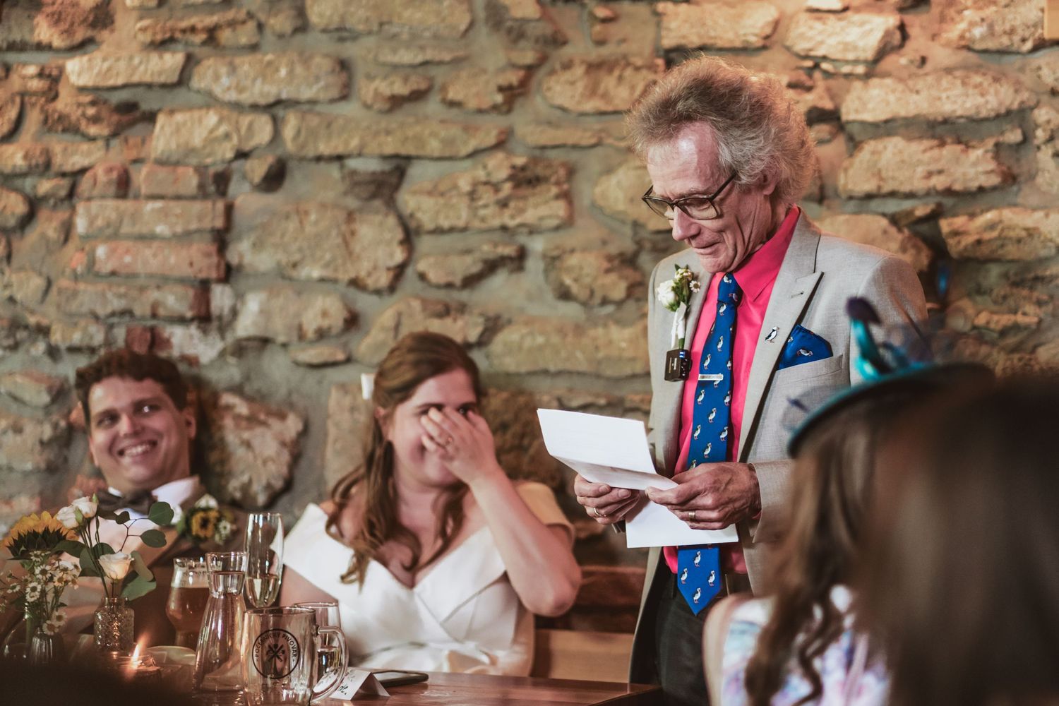 Elderly man in glasses and a gray suit reads a wedding speech while a bride covers her face laughing beside a smiling groom.