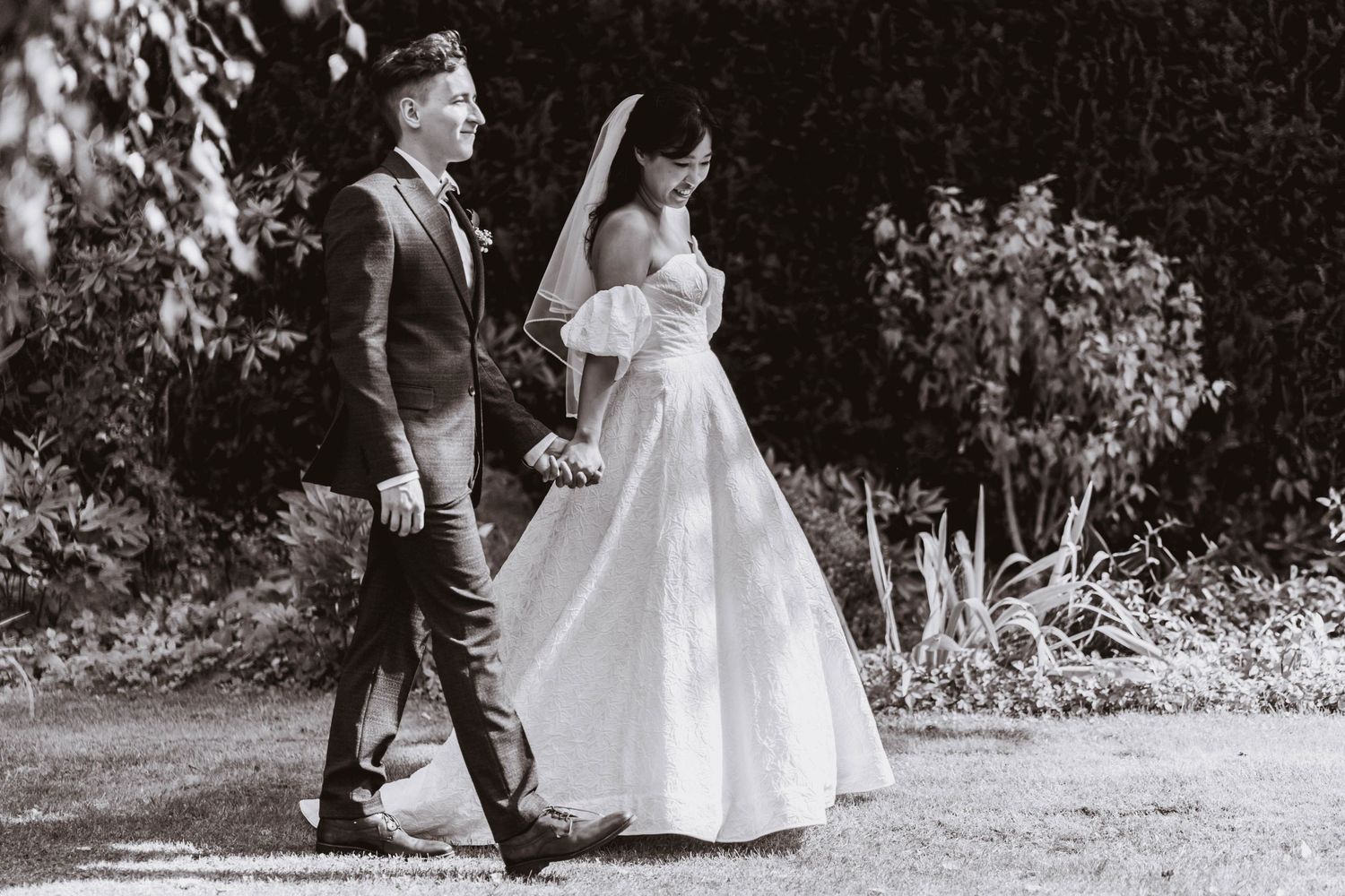 Black and white photo of a bride in a strapless gown and veil holding hands with a groom in a suit as they walk through a sunlit garden.