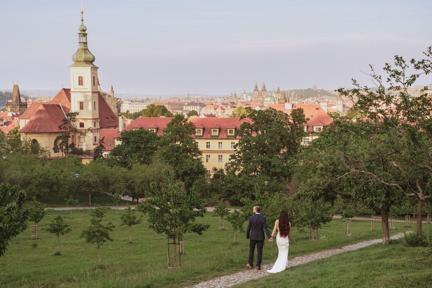 Bride in a white gown and groom in a dark suit walk hand in hand along a park path with a historic church tower and city rooftops behind.