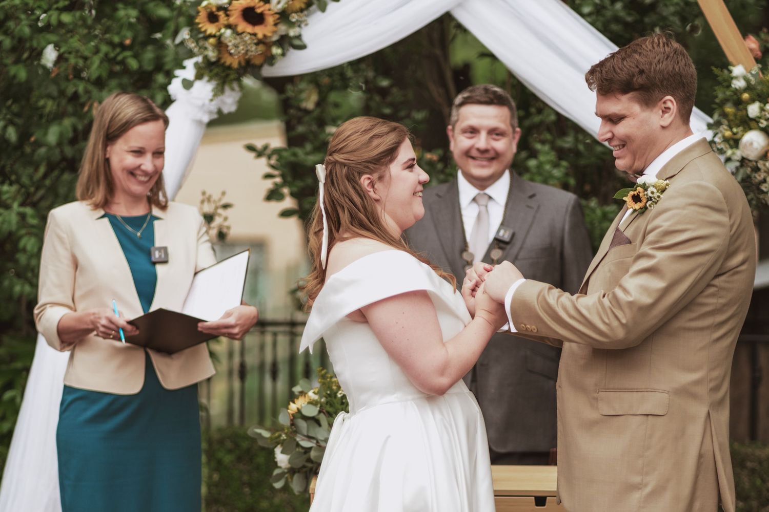Bride in a white gown and groom in a tan suit exchange vows during an outdoor wedding ceremony as the officiant and a guest look on.