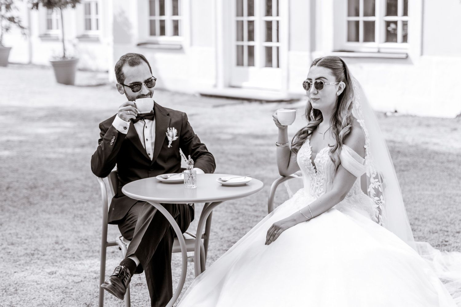 Bride and groom wearing sunglasses and sharing drinks at an outdoor table.