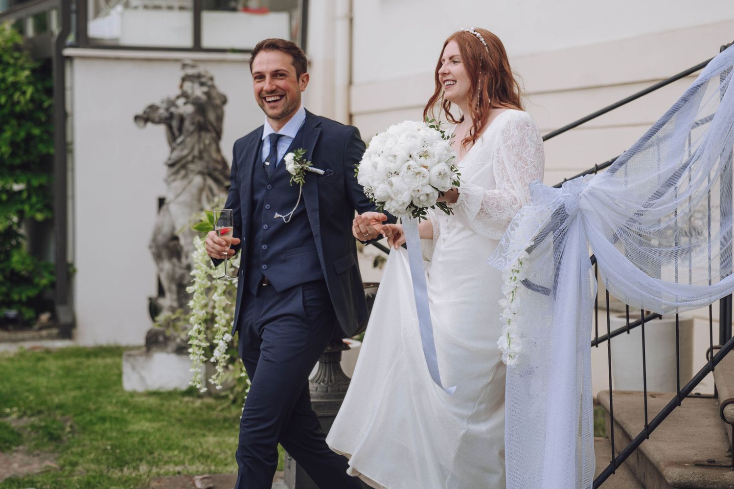 A smiling bride in a white gown holding a bouquet walks beside a groom in a navy suit with a boutonniere outside a stone building.