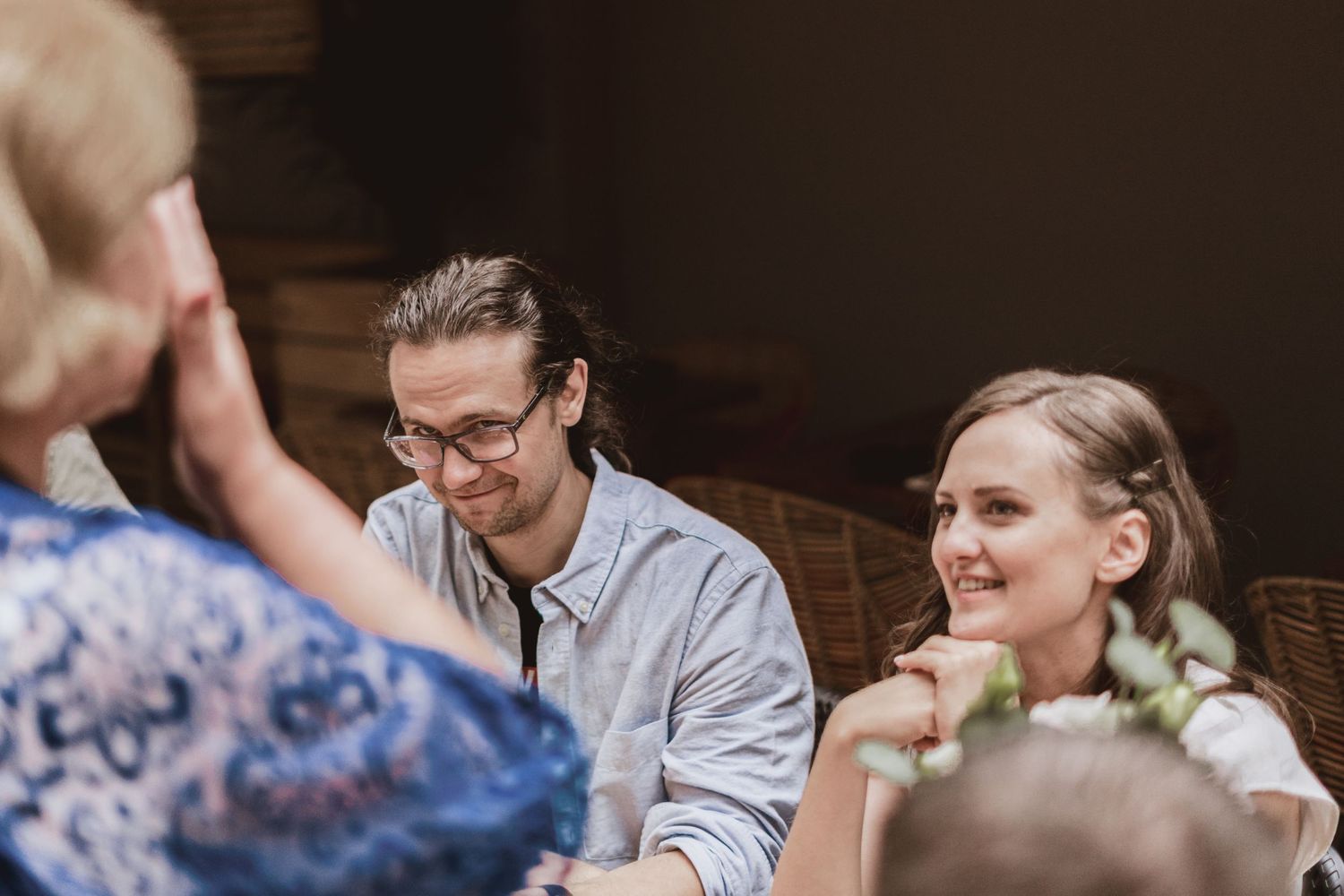Smiling woman with a floral hair wreath and a man in glasses laugh together at a gathering while a guest in lace shields her face.