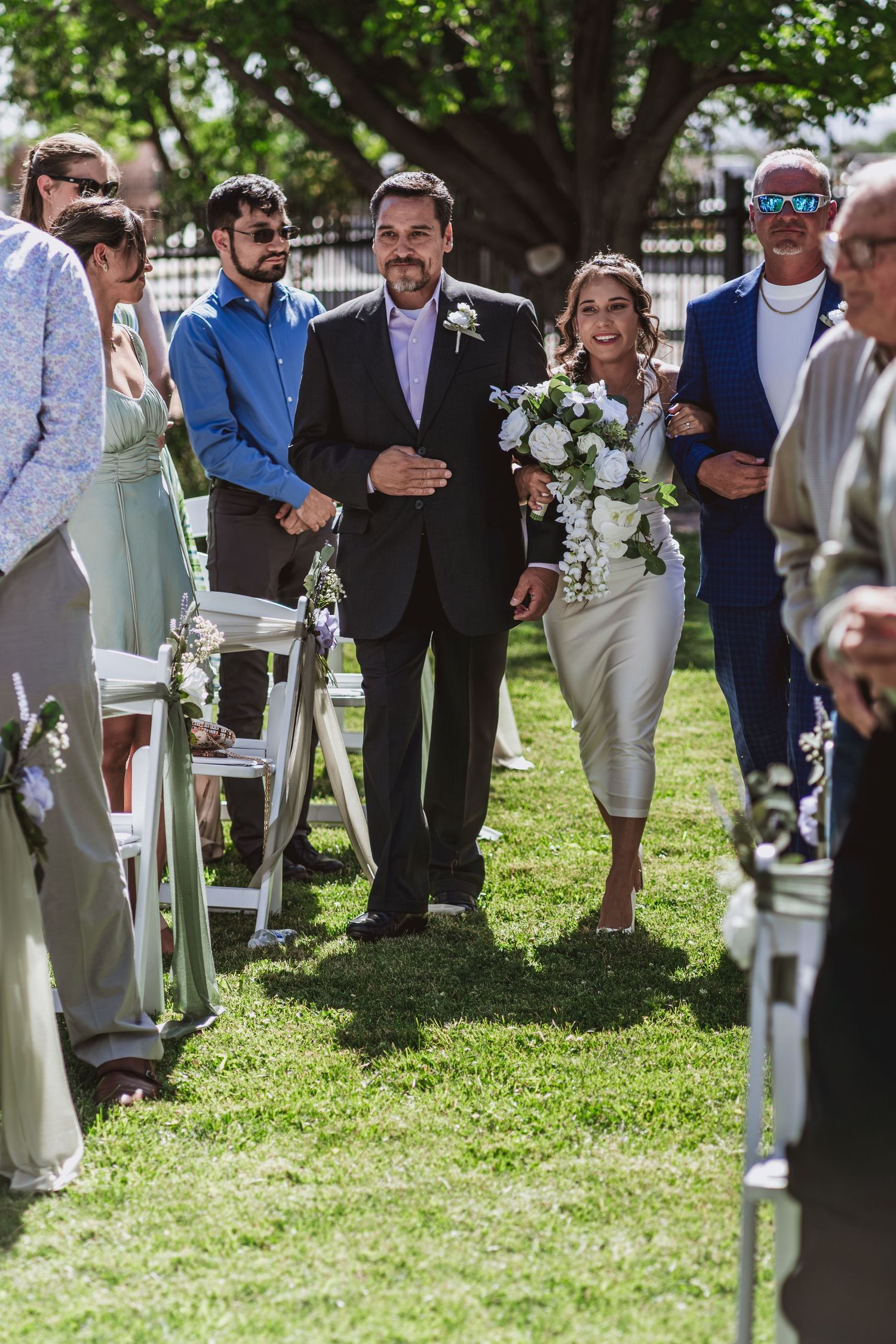 A father in a dark suit walks his smiling daughter, holding a white bouquet, down a grass aisle lined with white chairs and wedding guests.
