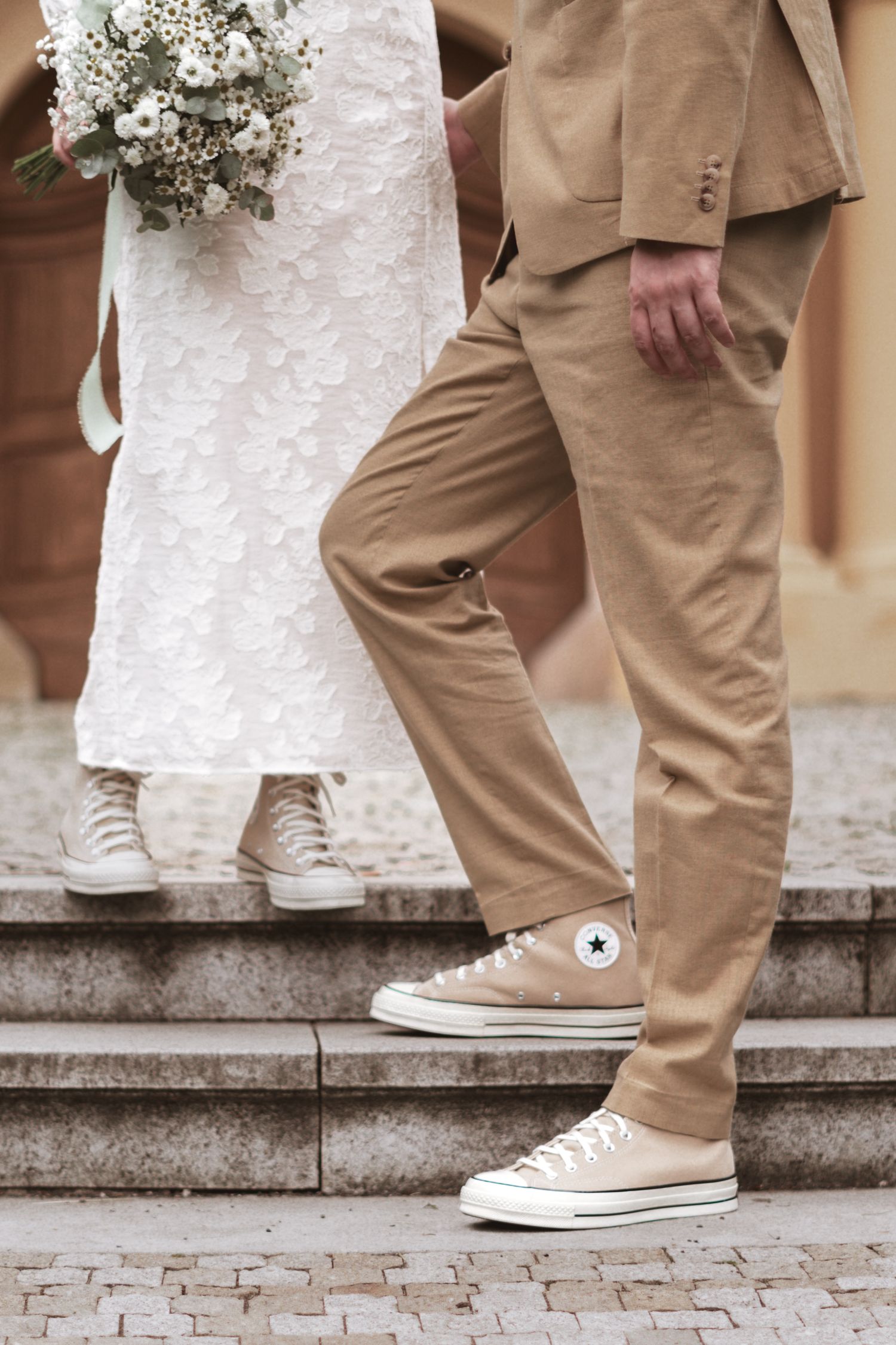 Bride in a white lace dress holding a bouquet stands beside a groom in a tan suit and beige Converse sneakers on stone steps.