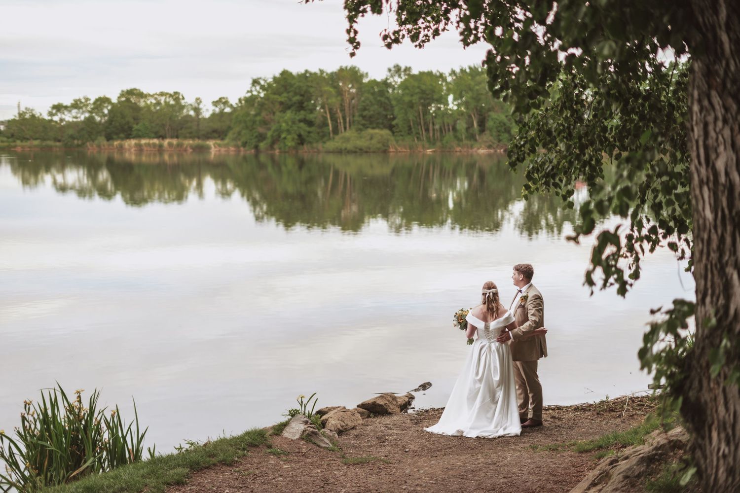 Bride in a white gown holding a bouquet stands beside her groom on a rocky shore overlooking a calm lake framed by a large tree.