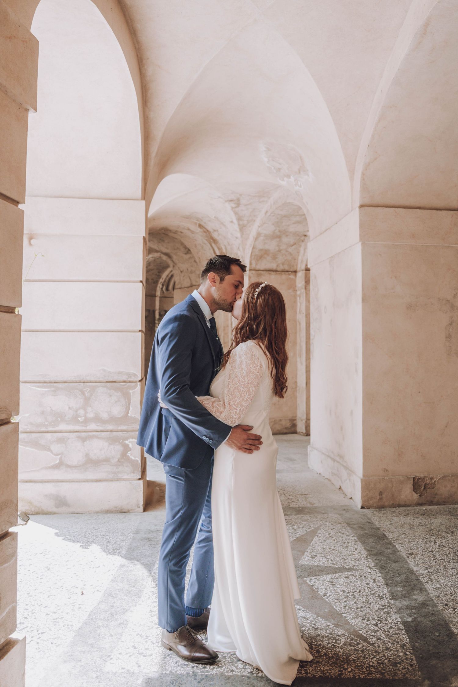 A bride in a long white dress and a groom in a blue suit share a kiss beneath the stone arches of a sunlit historic corridor.