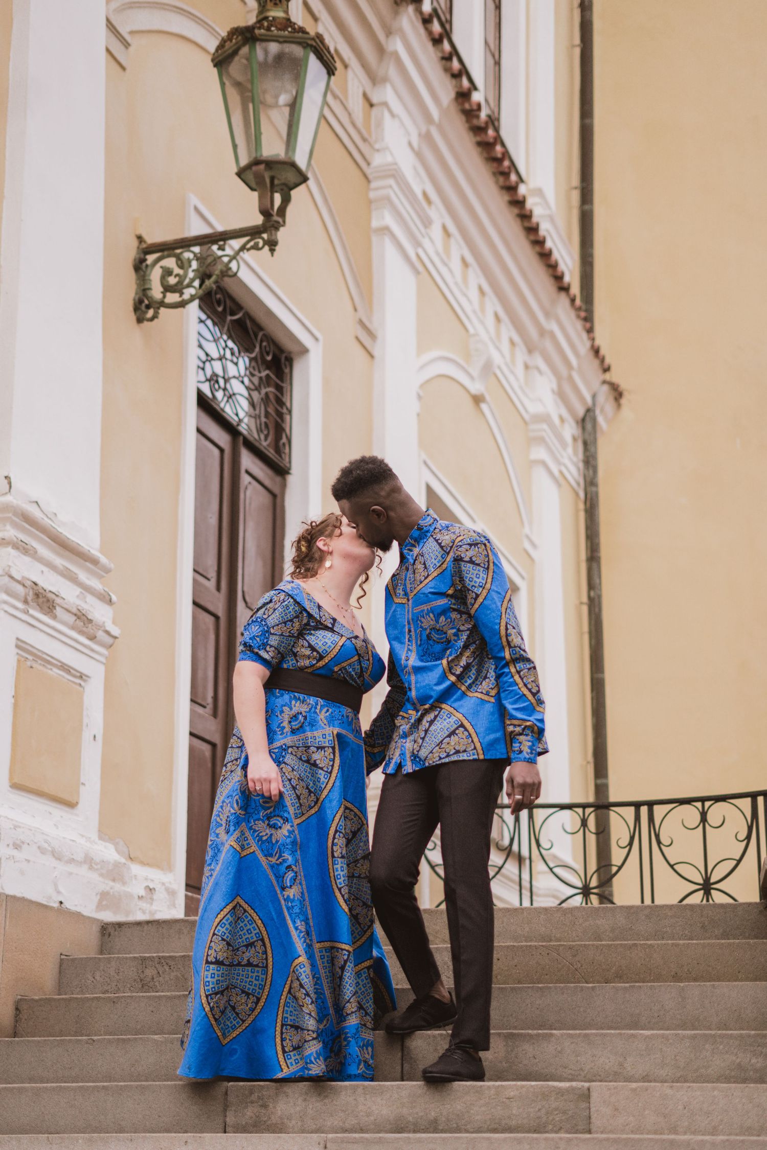 Couple in matching blue African wax print outfits share a kiss on stone steps outside a historic yellow building with a vintage lantern.