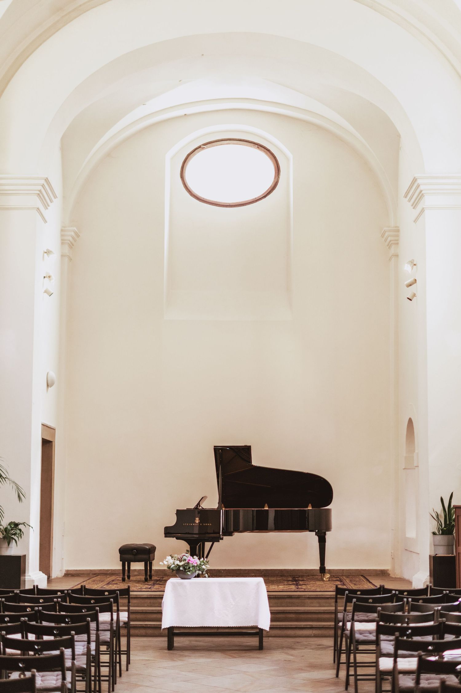 Black grand piano on a wooden stage beneath an oval window in a bright, arched chapel hall with rows of empty black chairs.