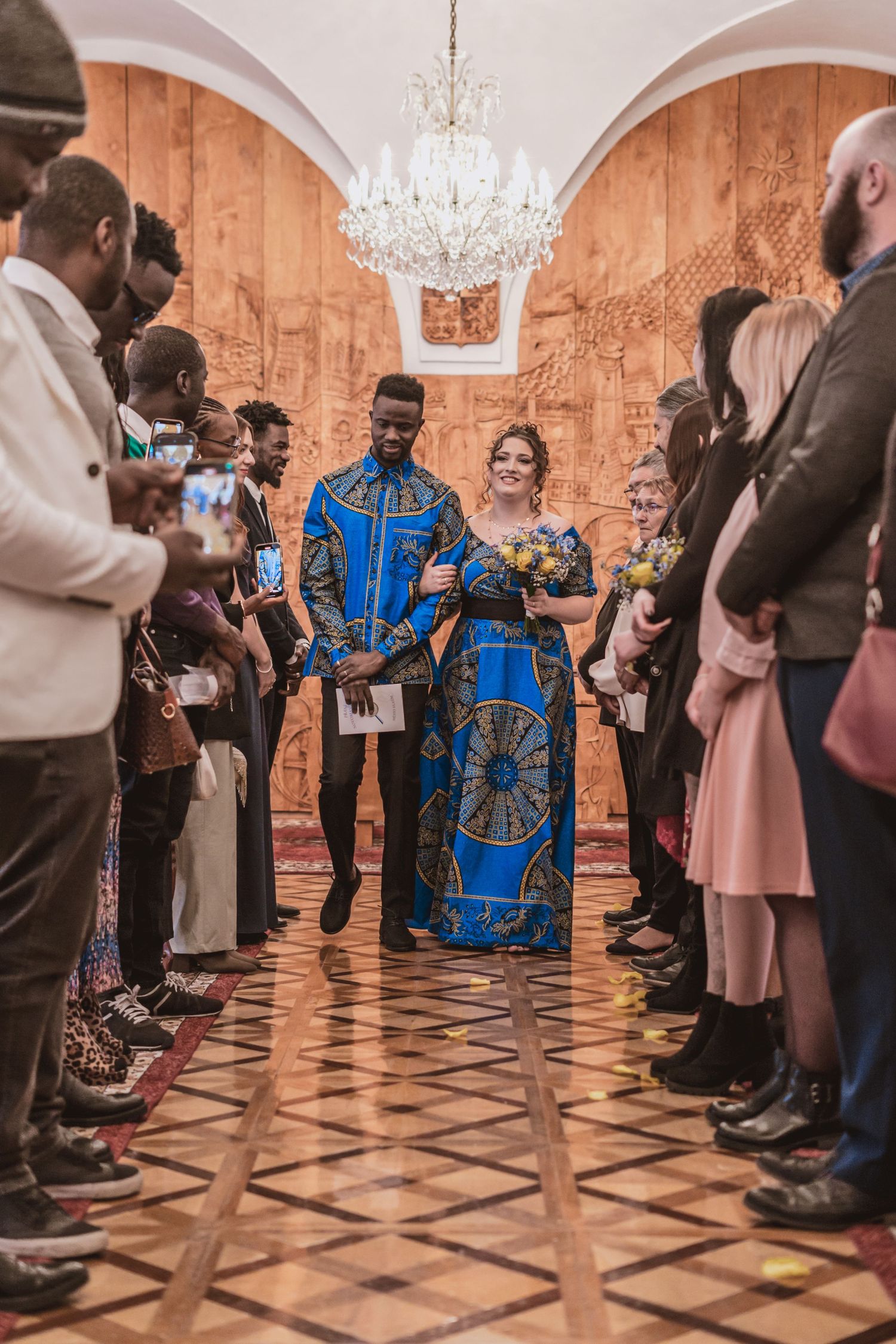 Bride and groom in matching blue African print outfits walk arm in arm down a wedding aisle past smiling guests under a crystal chandelier.