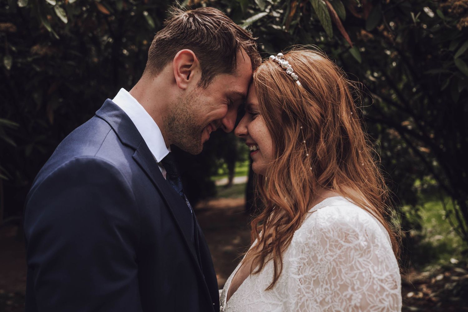 Smiling groom in a navy suit touches foreheads with a bride in a white lace dress and floral hair accessory in a leafy garden.