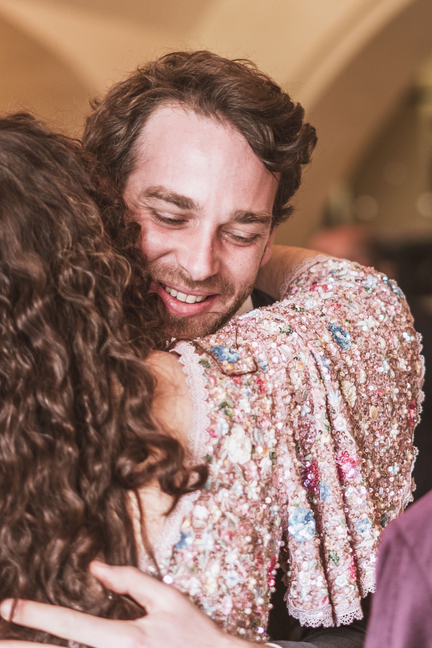 A smiling man with short brown hair warmly hugs a curly-haired person in a sparkly, sequined pink floral jacket at a festive gathering.