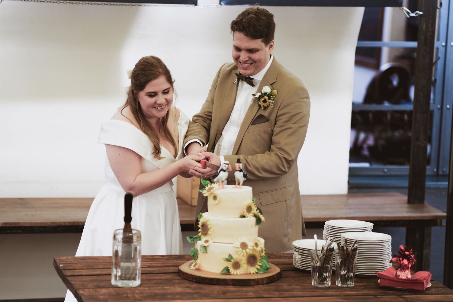 Smiling bride and groom together cut their sunflower-decorated three-tier wedding cake at a rustic reception table.