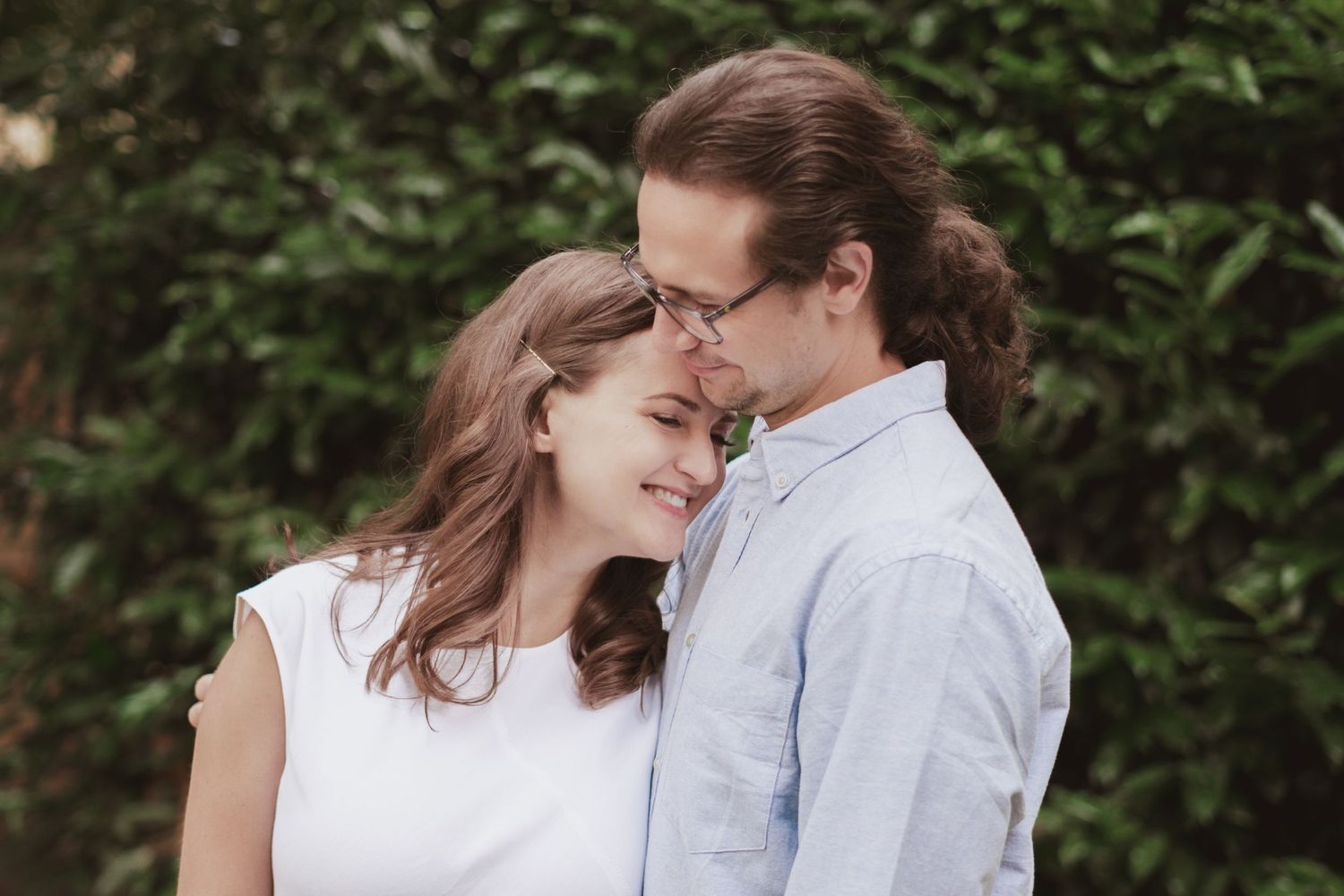 Smiling couple embracing outdoors as a man with long hair and glasses rests his forehead against a woman's head in front of green foliage.