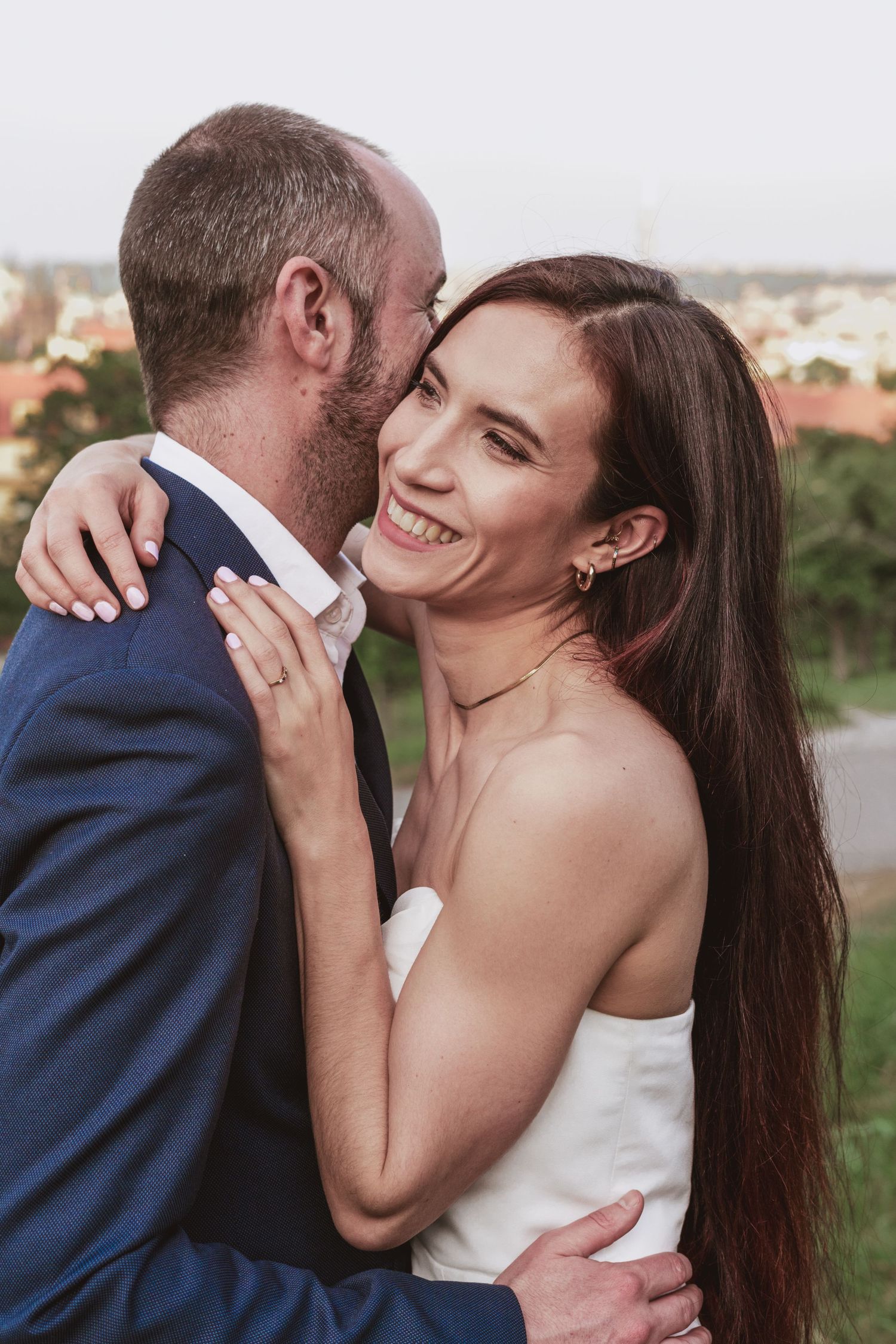 Smiling woman with long brown hair in a white strapless dress embraces a bearded man in a navy suit outdoors at sunset.