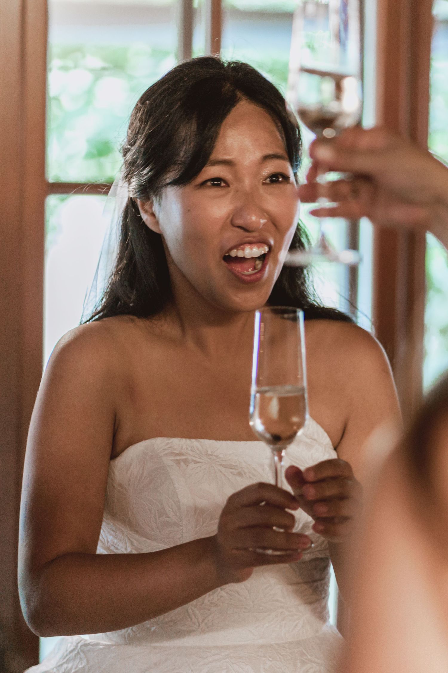 Smiling woman in a white strapless dress raises a champagne flute in a toast with another guest at a bright wedding celebration.
