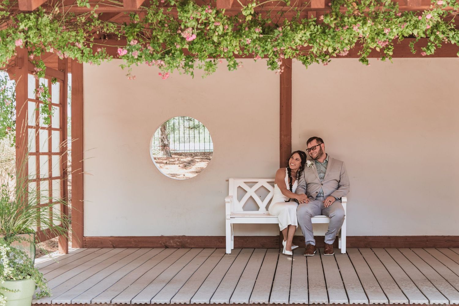 Couple sitting close together on a white bench on a wooden deck under a vine-covered pergola beside a wall with a round window.