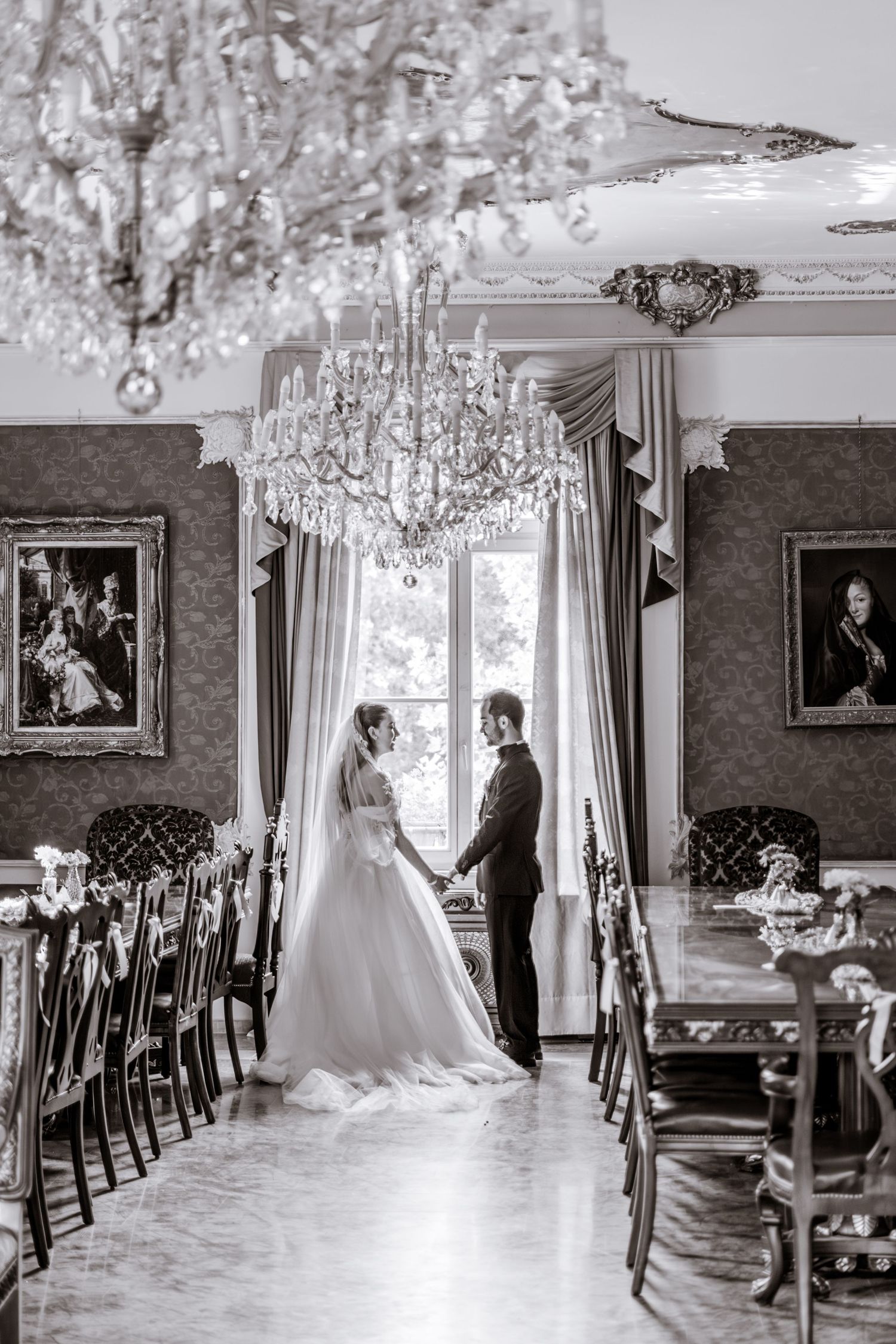 Bride and groom hold hands by a tall window in an elegant dining hall with crystal chandeliers, in black and white.