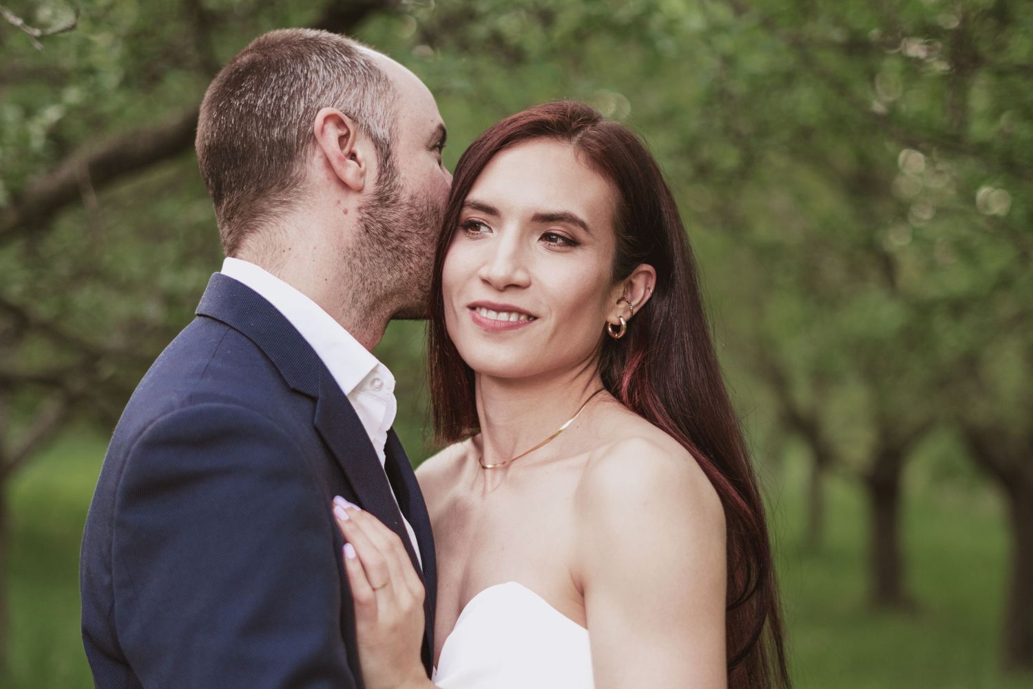 A bearded man in a navy suit kisses the cheek of a smiling woman with long dark hair in a white dress among green orchard trees.
