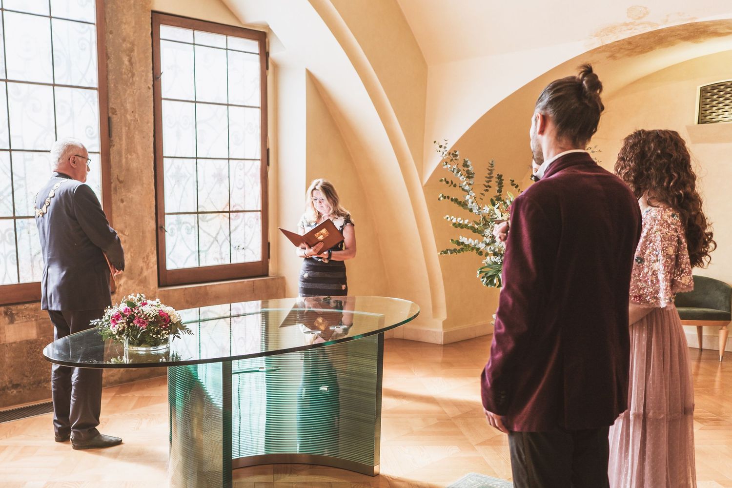 Couple in burgundy jacket and pink gown stand before an officiant at a glass table during a wedding ceremony in a bright arched room.