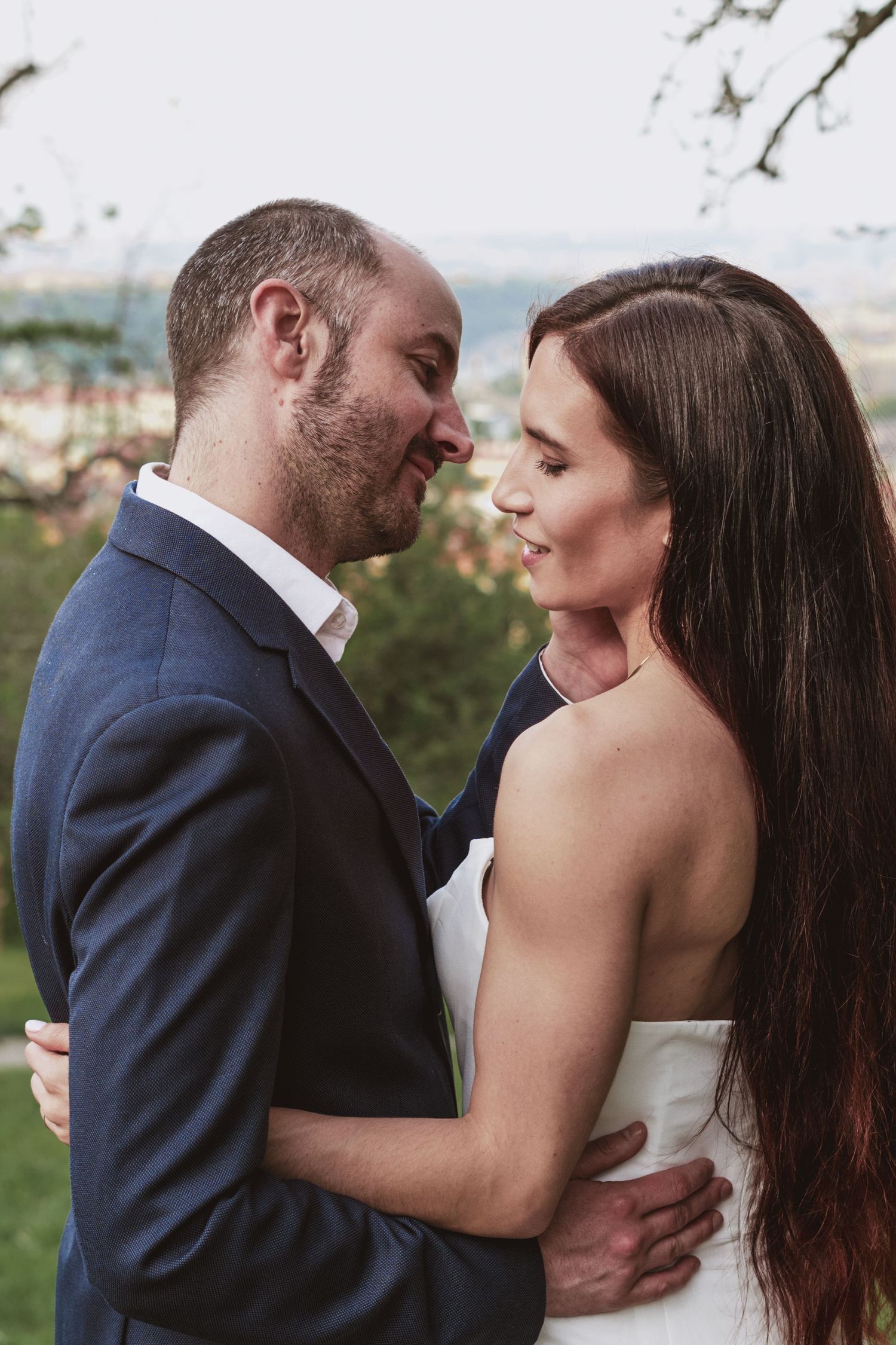 Smiling couple embracing outdoors at sunset, the man in a navy blazer and the woman in a white dress with long brown hair.