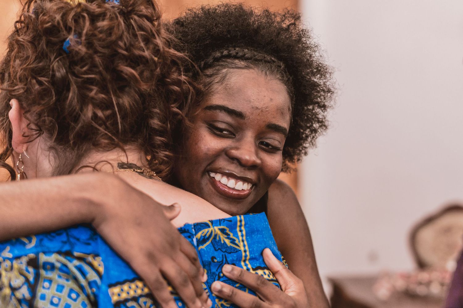 Two women share a warm, joyful hug as one smiles broadly, wearing a colorful patterned blue top in a bright indoor setting.